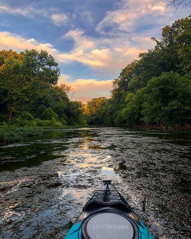 Front end of a kayak on a river. Trees line the sides of the river underneath a partly cloudy sky at the beginning of sunset.