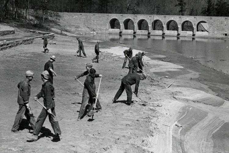 Black and white photo of peopel with the Civilian Conservation Corps working on a riverbank next to a dam.
