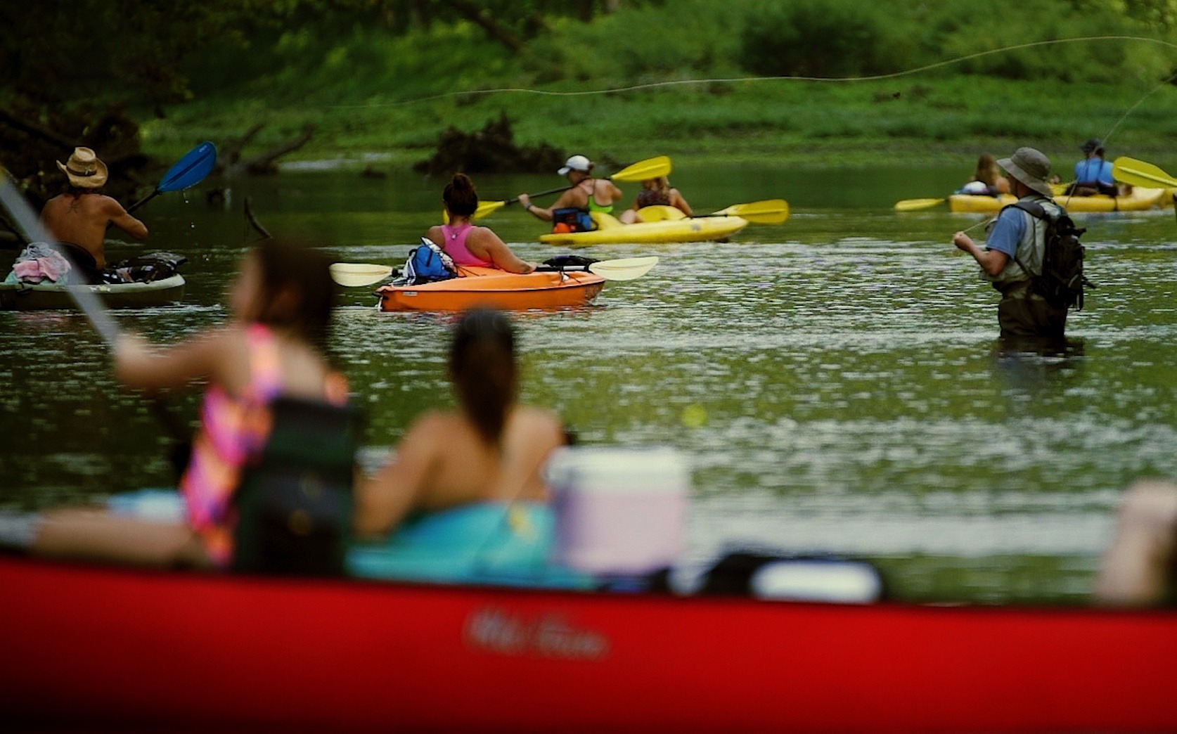 Anglers fishing on a river while kayakers paddle around them.