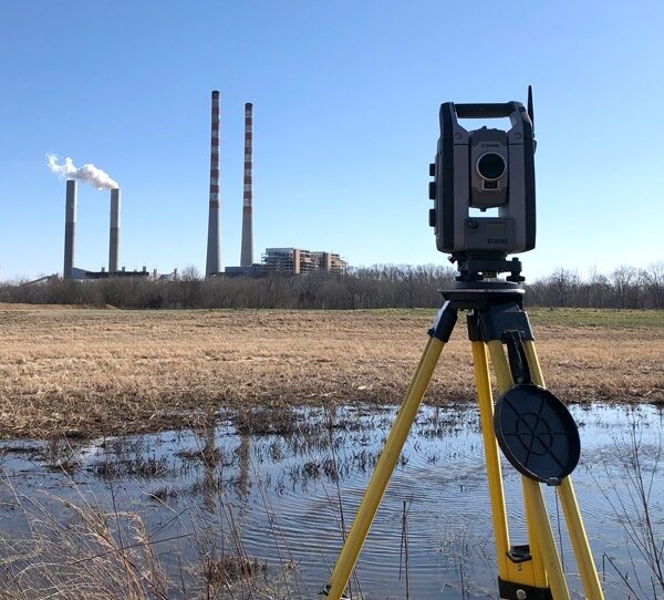 Surveying equipment set up at a wetland. In front of the equipment is a large field saturated with water. On the opposite end of the field is a factory with smoke billowing from the smokestacks.